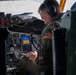 A U.S. military aviator is seated in the cockpit of a large military aircraft, reviewing a tablet device while in flight. The pilot is wearing a flight suit with a U.S. flag patch, Bose aviation headset, and has access to an extensive array of analog and digital cockpit instruments. The cockpit configuration and instrument panel suggest this is likely a large strategic aircraft such as a B-52 or KC-135.