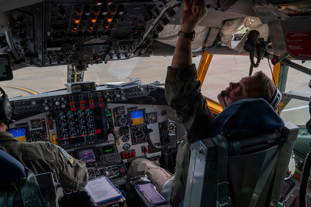 The image shows the interior cockpit of a large military aircraft, likely a Boeing B-52 Stratofortress or similar heavy bomber/transport, with two crew members conducting pre-flight or in-flight procedures. The right-seat pilot is reaching up to manipulate overhead panel switches while holding a communication device, while the left-seat crew member reviews documents. The cockpit displays a complex array of analog and digital instrumentation panels.