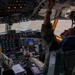 The image shows the interior cockpit of a large military aircraft, likely a Boeing B-52 Stratofortress or similar heavy bomber/transport, with two crew members conducting pre-flight or in-flight procedures. The right-seat pilot is reaching up to manipulate overhead panel switches while holding a communication device, while the left-seat crew member reviews documents. The cockpit displays a complex array of analog and digital instrumentation panels.