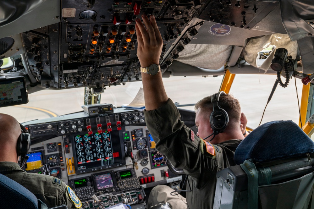 Two military aircrew members are seated in the cockpit of a large military aircraft, likely a KC-135 or similar tanker/transport aircraft. One crew member is reaching up to manipulate overhead panel switches while the other monitors instruments. Both are wearing flight suits, headsets, and military patches including visible US flag patches.