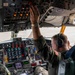 Two military aircrew members are seated in the cockpit of a large military aircraft, likely a KC-135 or similar tanker/transport aircraft. One crew member is reaching up to manipulate overhead panel switches while the other monitors instruments. Both are wearing flight suits, headsets, and military patches including visible US flag patches.