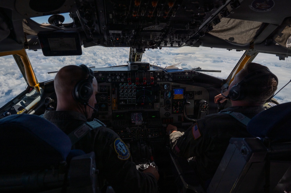 Two military aviators are seated in the cockpit of a large military aircraft, likely a KC-135 or similar tanker/transport, flying above a cloud layer. Both crew members are wearing flight suits, headsets, and communication equipment. The cockpit features an extensive analog and digital instrument panel typical of older-generation military aircraft.
