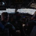 Two military aviators are seated in the cockpit of a large military aircraft, likely a KC-135 or similar tanker/transport, flying above a cloud layer. Both crew members are wearing flight suits, headsets, and communication equipment. The cockpit features an extensive analog and digital instrument panel typical of older-generation military aircraft.