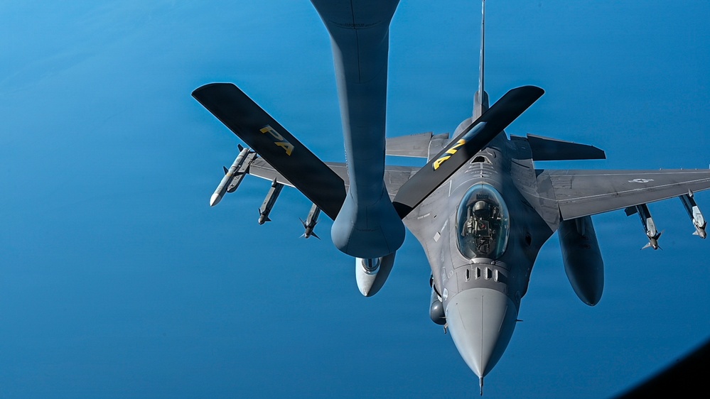 An F-16 Fighting Falcon jet fighter is captured from above during an aerial refueling operation, with a tanker aircraft's refueling boom extending toward the fighter. The aircraft is armed with visible air-to-air missiles on its wing pylons and carries a pilot in the single-seat cockpit. The image is taken from the perspective of the tanker aircraft looking down at the receiver jet against a clear blue sky.