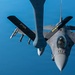 An F-16 Fighting Falcon jet fighter is captured from above during an aerial refueling operation, with a tanker aircraft's refueling boom extending toward the fighter. The aircraft is armed with visible air-to-air missiles on its wing pylons and carries a pilot in the single-seat cockpit. The image is taken from the perspective of the tanker aircraft looking down at the receiver jet against a clear blue sky.