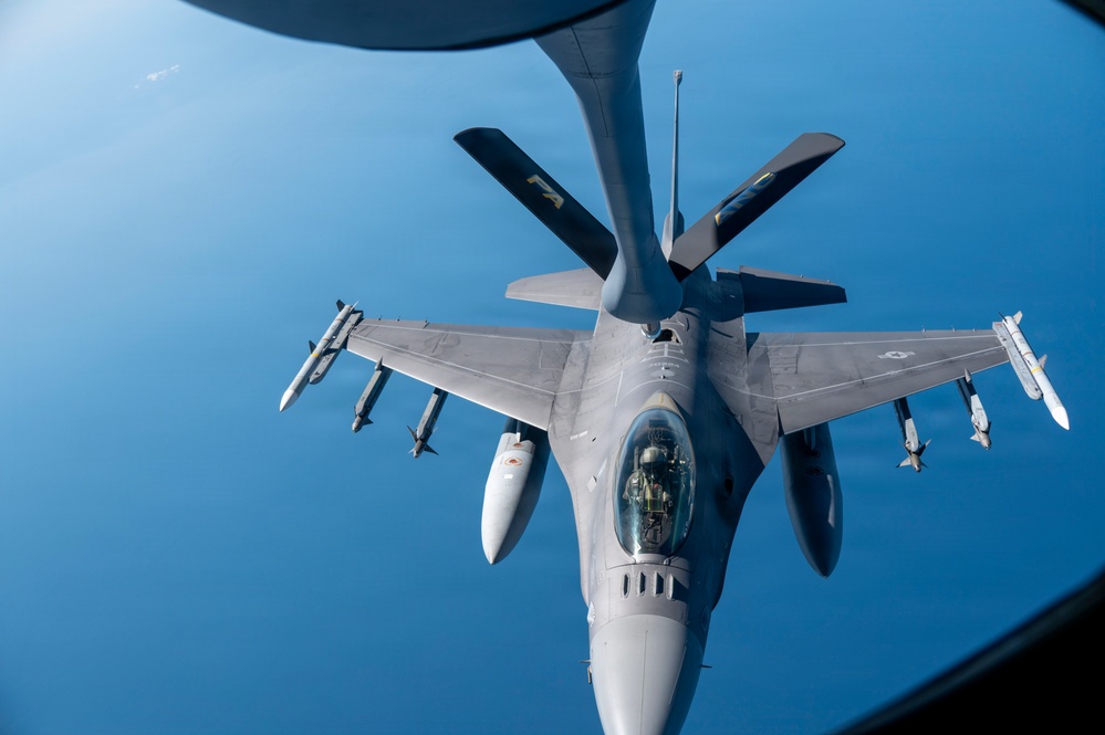 An F-16 Fighting Falcon fighter jet is captured from above during an aerial refueling operation with a tanker aircraft. The image is taken from the boom operator's position aboard the tanker, showing the refueling boom extending toward the F-16's receptacle. The aircraft is armed with multiple air-to-air missiles and carries external fuel tanks.