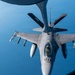 An F-16 Fighting Falcon fighter jet is captured from above during an aerial refueling operation with a tanker aircraft. The image is taken from the boom operator's position aboard the tanker, showing the refueling boom extending toward the F-16's receptacle. The aircraft is armed with multiple air-to-air missiles and carries external fuel tanks.