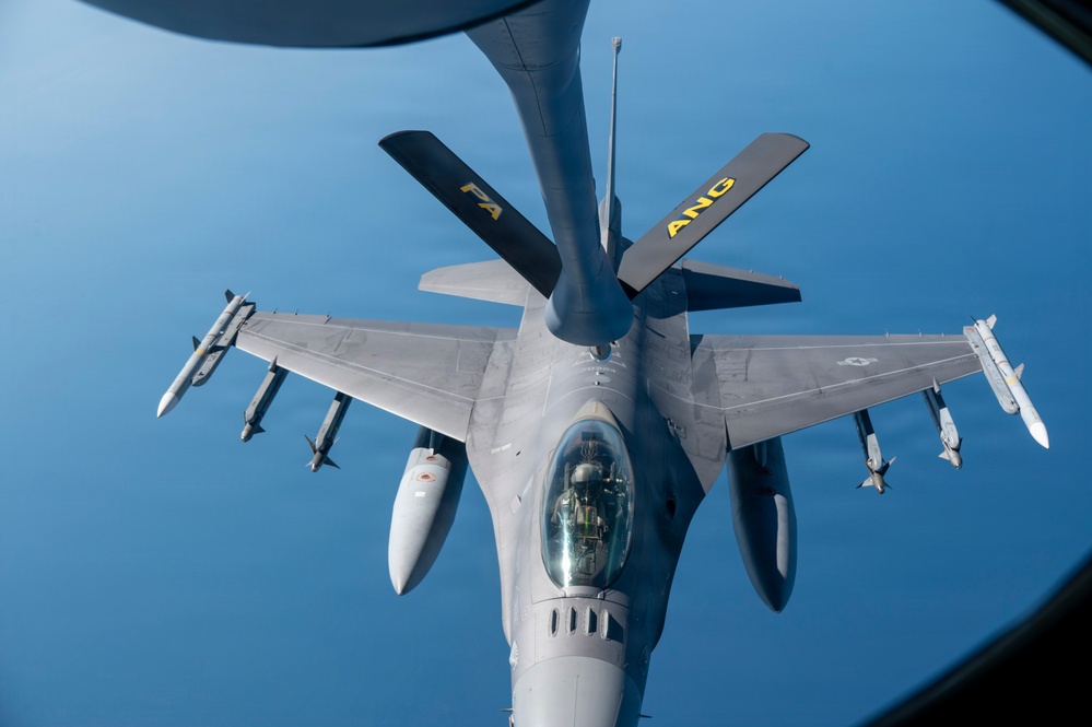 An aerial refueling operation is underway, with a KC-135 or similar tanker aircraft extending its boom toward an F-16 Fighting Falcon fighter jet. The photo is taken from the tanker's boom operator window looking down at the receiver aircraft. The F-16 is armed with multiple air-to-air missiles and carries external fuel tanks, and bears markings indicating it belongs to an Air National Guard (ANG) unit.