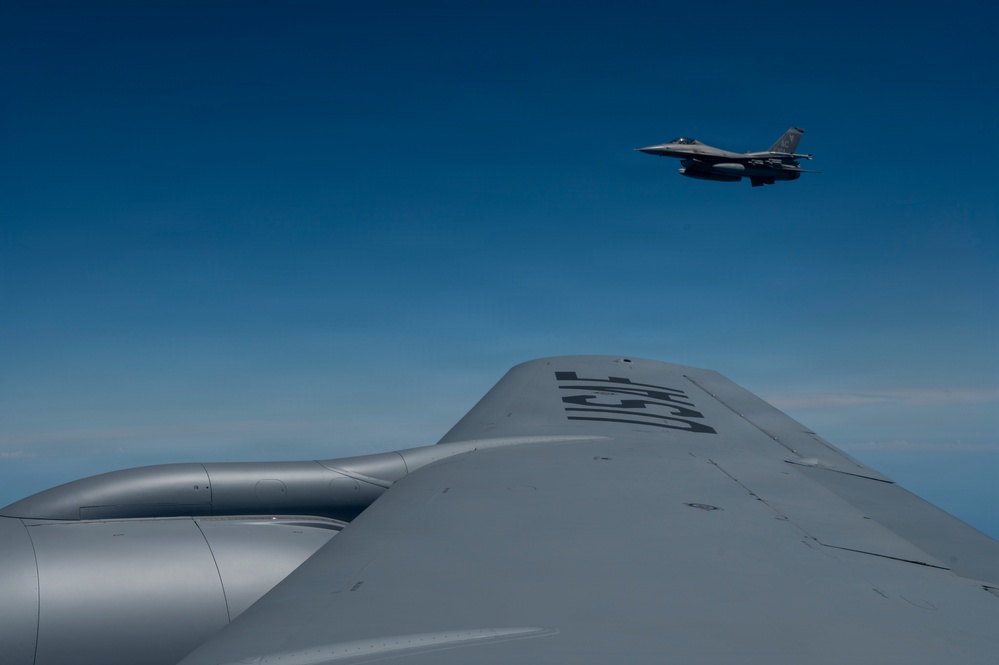 An aerial photograph taken from the fuselage or wing of a large military aircraft, likely a tanker or bomber, showing a fighter jet flying in formation above and behind. The foreground aircraft displays partial markings on its upper surface. The sky is clear and blue with minimal cloud cover at the horizon.