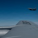 An aerial photograph taken from the fuselage or wing of a large military aircraft, likely a tanker or bomber, showing a fighter jet flying in formation above and behind. The foreground aircraft displays partial markings on its upper surface. The sky is clear and blue with minimal cloud cover at the horizon.