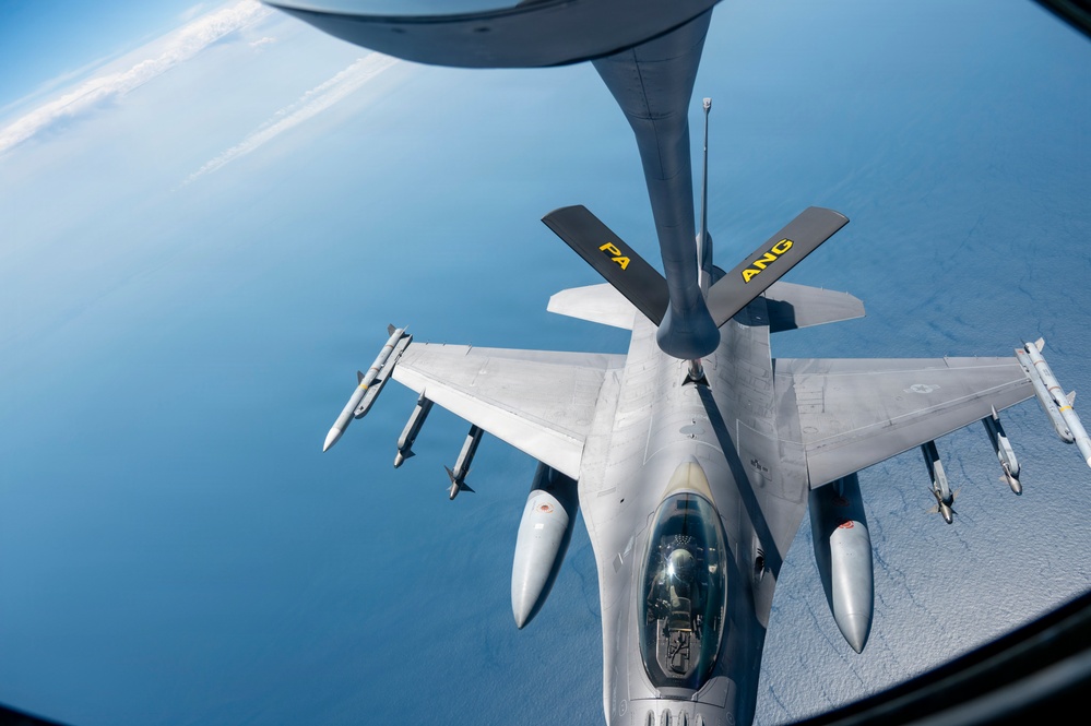 An aerial refueling operation is taking place between a tanker aircraft and an F-16 Fighting Falcon fighter jet at high altitude over a body of water. The tanker's refueling boom is extended and connected to the fighter, which is carrying multiple air-to-air missiles and other ordnance. The tanker is marked with 'PA ANG' indicating it belongs to the Pennsylvania Air National Guard.