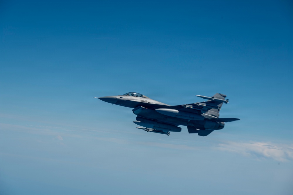 A single-seat fighter jet, consistent with an F-16 Fighting Falcon, is photographed in flight against a clear blue sky. The aircraft is armed with visible air-to-air missiles and carries external fuel tanks or munitions under its wings and fuselage. The image is taken from another aircraft at a similar altitude, providing a close lateral view.