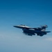 A single-seat fighter jet, consistent with an F-16 Fighting Falcon, is photographed in flight against a clear blue sky. The aircraft is armed with visible air-to-air missiles and carries external fuel tanks or munitions under its wings and fuselage. The image is taken from another aircraft at a similar altitude, providing a close lateral view.