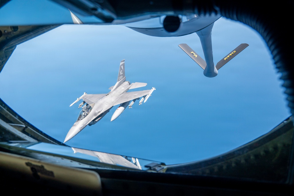 An aerial refueling operation is captured from the boom operator's station of a tanker aircraft, showing an F-16 Fighting Falcon approaching for fuel transfer. The photo is taken through the rear observation window of what appears to be a KC-135 Stratotanker, with the refueling boom visible in the upper portion of the frame. The F-16 bears markings consistent with the Air National Guard (ANG), as indicated by text visible on the tanker's boom.