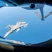 An aerial refueling operation is captured from the boom operator's station of a tanker aircraft, showing an F-16 Fighting Falcon approaching for fuel transfer. The photo is taken through the rear observation window of what appears to be a KC-135 Stratotanker, with the refueling boom visible in the upper portion of the frame. The F-16 bears markings consistent with the Air National Guard (ANG), as indicated by text visible on the tanker's boom.