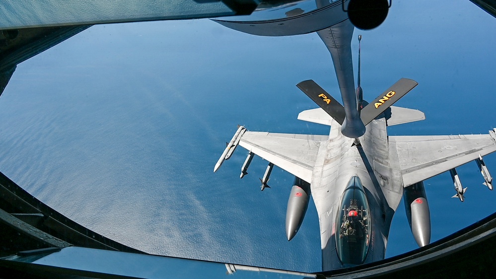 An aerial refueling operation is captured from the boom operator's position aboard a tanker aircraft, showing an F-16 Fighting Falcon approaching to receive fuel. The receiver aircraft bears 'PA' and 'ANG' markings, identifying it as belonging to the Pennsylvania Air National Guard. The scene is photographed over a large body of open water visible far below.
