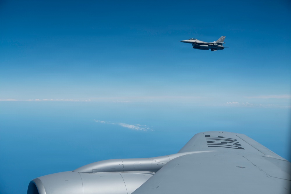 An aerial photograph taken from aboard a large military aircraft, likely a tanker such as a KC-135 or KC-10, showing a fighter jet — identifiable as an F-16 Fighting Falcon — flying in formation at higher altitude. The foreground shows the upper fuselage and engine nacelle of the tanker aircraft with partially visible tail numbers. The scene takes place over open ocean or sea under clear blue skies with a thin cloud layer visible on the horizon.