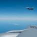 An aerial photograph taken from aboard a large military aircraft, likely a tanker such as a KC-135 or KC-10, showing a fighter jet — identifiable as an F-16 Fighting Falcon — flying in formation at higher altitude. The foreground shows the upper fuselage and engine nacelle of the tanker aircraft with partially visible tail numbers. The scene takes place over open ocean or sea under clear blue skies with a thin cloud layer visible on the horizon.