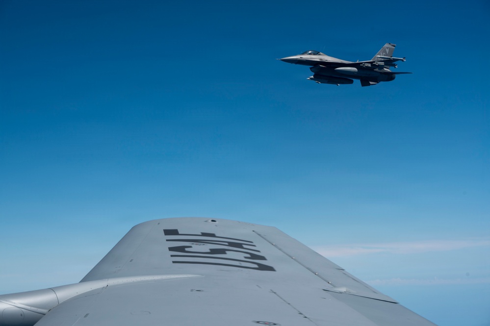 An aerial photograph taken from the fuselage or wing of a large military aircraft, showing an F-16 Fighting Falcon flying overhead against a clear blue sky. The foreground aircraft appears to be a tanker or large transport, with partial markings visible on its upper fuselage. The image captures a mid-air formation or refueling proximity scenario.