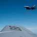 An aerial photograph taken from the fuselage or wing of a large military aircraft, showing an F-16 Fighting Falcon flying overhead against a clear blue sky. The foreground aircraft appears to be a tanker or large transport, with partial markings visible on its upper fuselage. The image captures a mid-air formation or refueling proximity scenario.