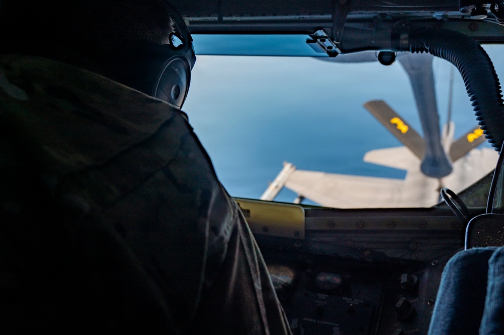 The image shows a view from inside a military aircraft cockpit, with a pilot or crew member visible in silhouette on the left side. Through the windscreen, another aircraft is visible in close proximity, suggesting an aerial refueling operation is taking place. The scene is set at altitude with a blue sky background.