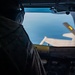 The image shows a view from inside a military aircraft cockpit, with a pilot or crew member visible in silhouette on the left side. Through the windscreen, another aircraft is visible in close proximity, suggesting an aerial refueling operation is taking place. The scene is set at altitude with a blue sky background.