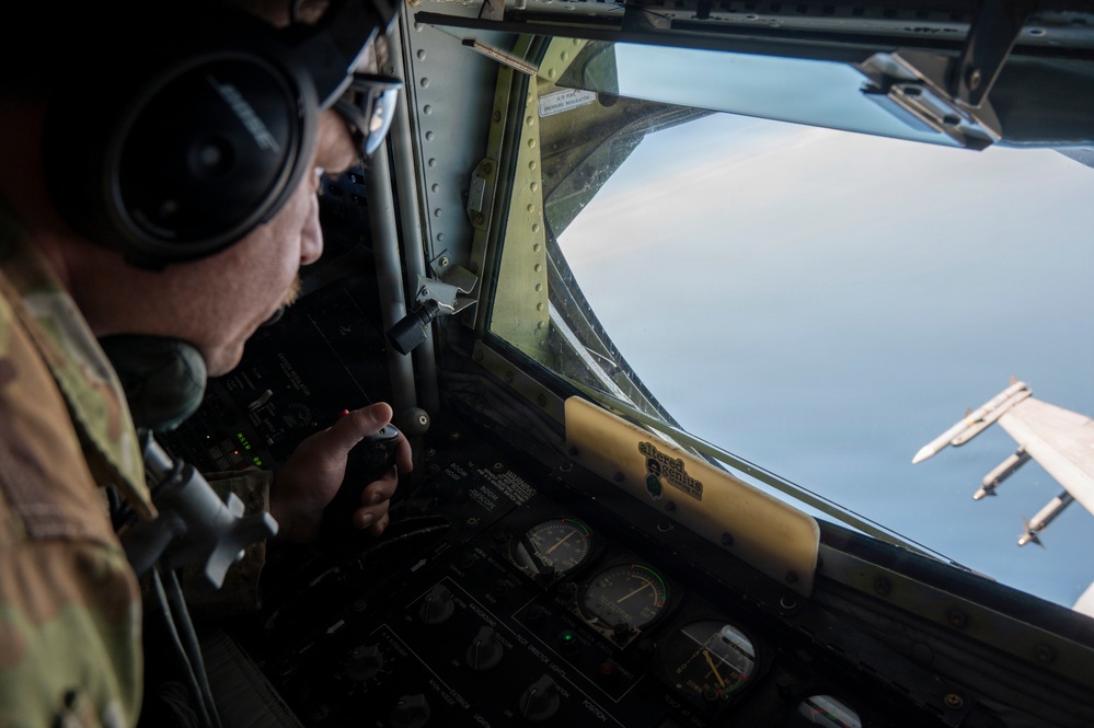 A military aircrew member wearing a flight helmet and headset is seated at a cockpit station, operating controls while in flight. Through the cockpit window, another military aircraft is visible flying in close proximity, suggesting a formation flight or aerial refueling operation. The cockpit displays analog gauges and instrumentation consistent with a large military transport or tanker aircraft.