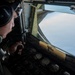 A military aircrew member wearing a flight helmet and headset is seated at a cockpit station, operating controls while in flight. Through the cockpit window, another military aircraft is visible flying in close proximity, suggesting a formation flight or aerial refueling operation. The cockpit displays analog gauges and instrumentation consistent with a large military transport or tanker aircraft.
