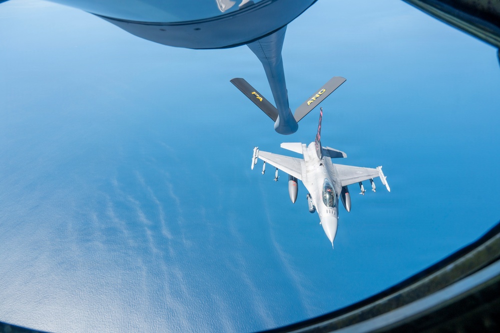 An aerial refueling operation is captured from the boom operator's window of a tanker aircraft, showing an F-16 Fighting Falcon approaching the refueling boom. The tanker's boom is extended downward toward the fighter jet, with 'PA ANG' (Pennsylvania Air National Guard) markings visible on the boom's stabilizing fins. The scene is set over open water or ocean at high altitude.