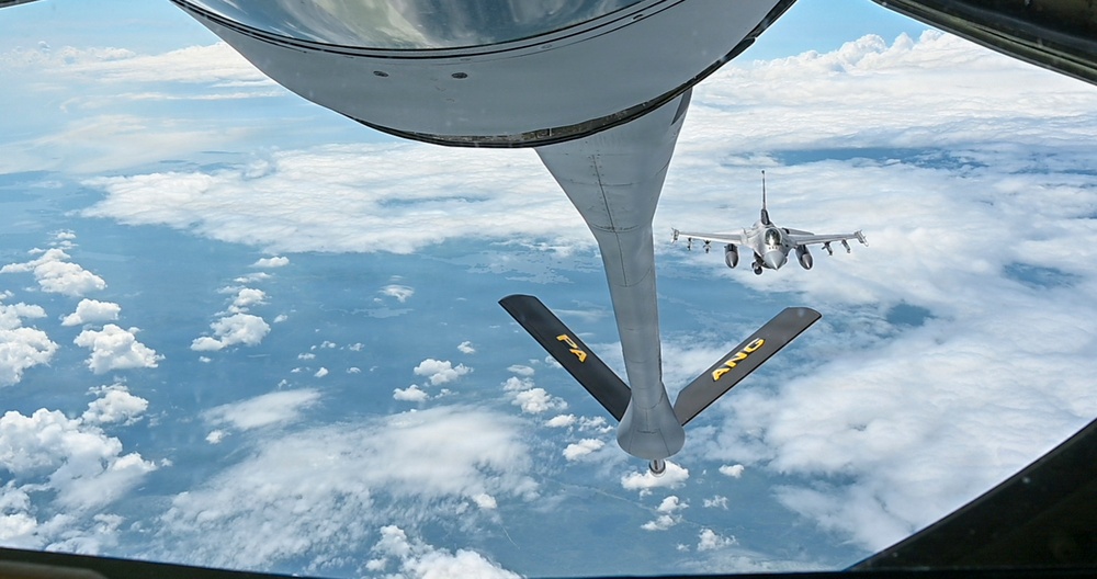 An aerial refueling operation is captured from the boom operator's position aboard a tanker aircraft, showing the refueling boom extended downward with stabilizing ruddervators marked 'PA' and 'ANG'. An F-16 Fighting Falcon fighter jet is visible in the background, approaching or awaiting its turn to receive fuel. The scene is set at altitude above a cloud-covered landscape under blue skies.