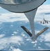 An aerial refueling operation is captured from the boom operator's position aboard a tanker aircraft, showing the refueling boom extended downward with stabilizing ruddervators marked 'PA' and 'ANG'. An F-16 Fighting Falcon fighter jet is visible in the background, approaching or awaiting its turn to receive fuel. The scene is set at altitude above a cloud-covered landscape under blue skies.