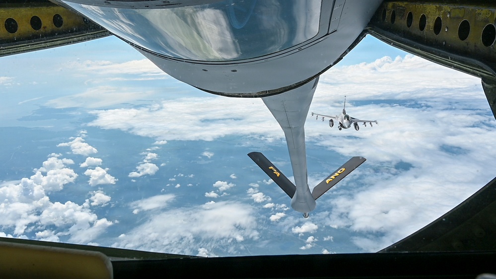 This image is taken from the boom operator's position inside a tanker aircraft, looking down through the rear observation window during an aerial refueling operation. The refueling boom extends downward with markings 'PA' and 'ANG' (Pennsylvania Air National Guard) visible on its ruddervators. A fighter jet, consistent with an F-16 Fighting Falcon, is visible in the background awaiting its turn or having just completed refueling.