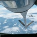 This image is taken from the boom operator's position inside a tanker aircraft, looking down through the rear observation window during an aerial refueling operation. The refueling boom extends downward with markings 'PA' and 'ANG' (Pennsylvania Air National Guard) visible on its ruddervators. A fighter jet, consistent with an F-16 Fighting Falcon, is visible in the background awaiting its turn or having just completed refueling.