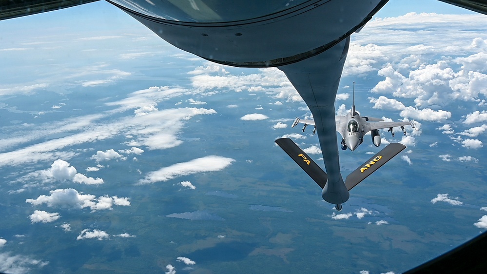 An aerial refueling operation is captured from the perspective of a tanker aircraft, showing its refueling boom extended toward an approaching F-16 Fighting Falcon fighter jet. The tanker's boom stabilizer fins are clearly marked with 'FA' and 'ANG', indicating Air National Guard affiliation. The scene takes place at high altitude over a landscape partially covered by scattered cumulus clouds.