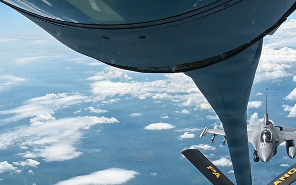 An aerial refueling operation is captured from the perspective of a tanker aircraft, showing its refueling boom extended toward an approaching F-16 Fighting Falcon fighter jet. The tanker's boom stabilizer fins are clearly marked with 'FA' and 'ANG', indicating Air National Guard affiliation. The scene takes place at high altitude over a landscape partially covered by scattered cumulus clouds.
