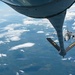 An aerial refueling operation is captured from the perspective of a tanker aircraft, showing its refueling boom extended toward an approaching F-16 Fighting Falcon fighter jet. The tanker's boom stabilizer fins are clearly marked with 'FA' and 'ANG', indicating Air National Guard affiliation. The scene takes place at high altitude over a landscape partially covered by scattered cumulus clouds.
