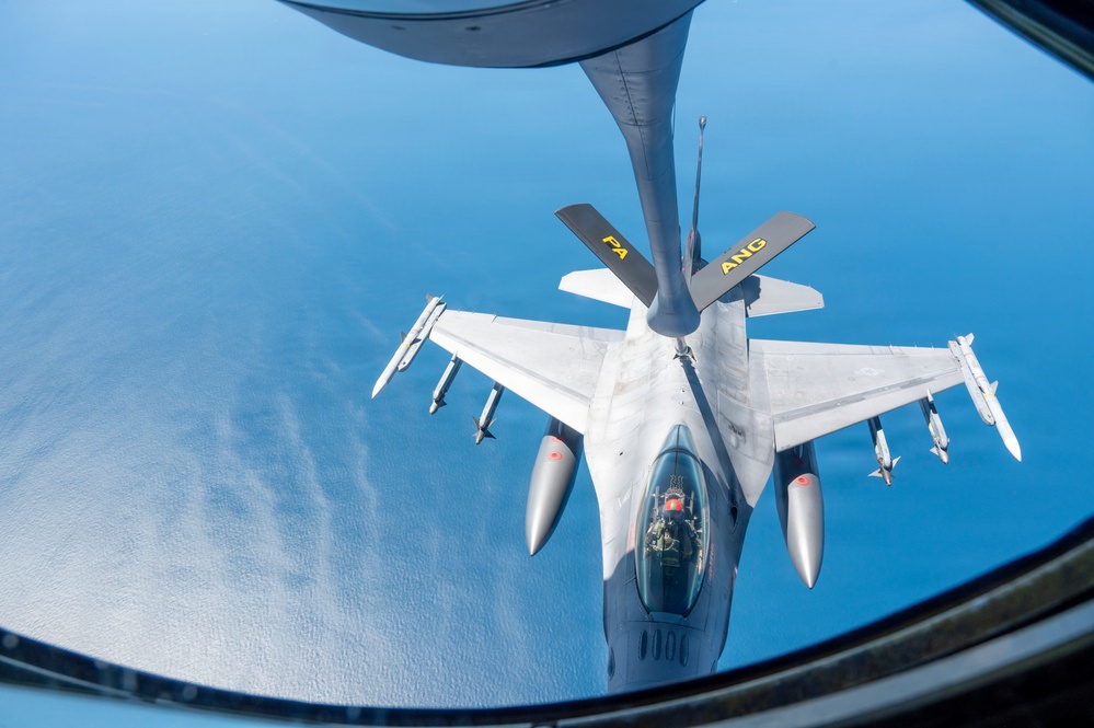 An aerial refueling operation is captured from the perspective of a tanker aircraft, showing an F-16 Fighting Falcon receiving fuel via a boom refueling system over open water. The fighter jet bears markings indicating it belongs to the Air National Guard (ANG) with a 'PA' designation, suggesting Pennsylvania Air National Guard. The aircraft is armed with multiple air-to-air missiles and external fuel tanks.