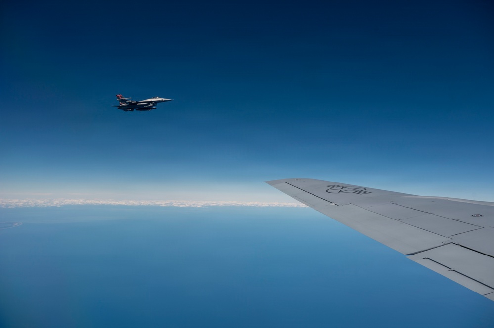 An aerial photograph taken from inside or alongside a large military aircraft, showing its wing in the foreground and a fighter jet flying in formation nearby. The scene is set at high altitude above a calm, reflective body of water under a clear deep blue sky. A thin layer of clouds is visible near the horizon.