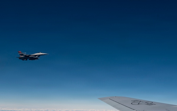 An aerial photograph taken from inside or alongside a large military aircraft, showing its wing in the foreground and a fighter jet flying in formation nearby. The scene is set at high altitude above a calm, reflective body of water under a clear deep blue sky. A thin layer of clouds is visible near the horizon.