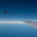 An aerial photograph taken from inside or alongside a large military aircraft, showing its wing in the foreground and a fighter jet flying in formation nearby. The scene is set at high altitude above a calm, reflective body of water under a clear deep blue sky. A thin layer of clouds is visible near the horizon.