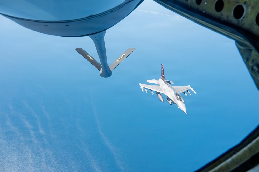 An aerial refueling operation is captured from the boom operator's window of a tanker aircraft, showing an F-16 Fighting Falcon approaching for fuel transfer. The refueling boom with 'PA' and 'ANG' markings (indicating Pennsylvania Air National Guard) is visible in the foreground. The scene takes place at high altitude over what appears to be a large body of water under clear blue skies.