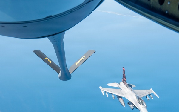 An aerial refueling operation is captured from the boom operator's window of a tanker aircraft, showing an F-16 Fighting Falcon approaching for fuel transfer. The refueling boom with 'PA' and 'ANG' markings (indicating Pennsylvania Air National Guard) is visible in the foreground. The scene takes place at high altitude over what appears to be a large body of water under clear blue skies.