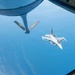 An aerial refueling operation is captured from the boom operator's window of a tanker aircraft, showing an F-16 Fighting Falcon approaching for fuel transfer. The refueling boom with 'PA' and 'ANG' markings (indicating Pennsylvania Air National Guard) is visible in the foreground. The scene takes place at high altitude over what appears to be a large body of water under clear blue skies.