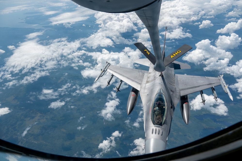 An aerial refueling operation is taking place between a tanker aircraft (visible at top) and an F-16 Fighting Falcon fighter jet. The F-16, marked with 'ANG' (Air National Guard) and 'FA' tail codes, is receiving fuel via a boom refueling system at high altitude. The image is taken from the tanker aircraft's boom operator window, looking down at the receiver aircraft with cloud cover and terrain visible far below.