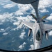 An aerial refueling operation is taking place between a tanker aircraft (visible at top) and an F-16 Fighting Falcon fighter jet. The F-16, marked with 'ANG' (Air National Guard) and 'FA' tail codes, is receiving fuel via a boom refueling system at high altitude. The image is taken from the tanker aircraft's boom operator window, looking down at the receiver aircraft with cloud cover and terrain visible far below.