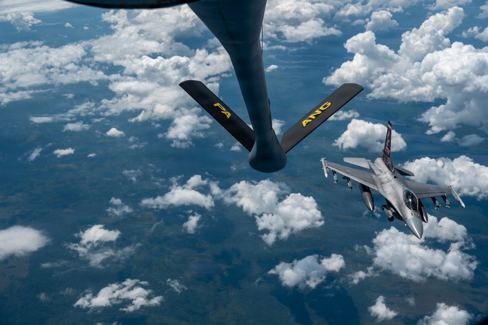 An aerial refueling operation is captured from the perspective of a tanker aircraft, showing its refueling boom extended toward an approaching F-16 Fighting Falcon fighter jet. The boom is marked with yellow letters 'FA' and 'ANG', indicating Air National Guard affiliation. The scene takes place at high altitude above scattered cumulus clouds with terrain visible far below.