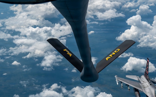 An aerial refueling operation is captured from the perspective of a tanker aircraft, showing its refueling boom extended toward an approaching F-16 Fighting Falcon fighter jet. The boom is marked with yellow letters 'FA' and 'ANG', indicating Air National Guard affiliation. The scene takes place at high altitude above scattered cumulus clouds with terrain visible far below.
