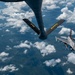 An aerial refueling operation is captured from the perspective of a tanker aircraft, showing its refueling boom extended toward an approaching F-16 Fighting Falcon fighter jet. The boom is marked with yellow letters 'FA' and 'ANG', indicating Air National Guard affiliation. The scene takes place at high altitude above scattered cumulus clouds with terrain visible far below.