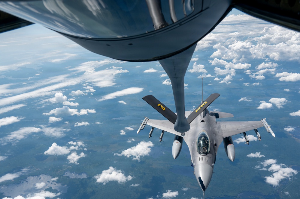 An aerial refueling operation is captured from the perspective of a tanker aircraft, showing its refueling boom extended toward an F-16 Fighting Falcon fighter jet. The F-16 is marked with 'PA' and 'ANG' in yellow lettering, indicating it belongs to the Pennsylvania Air National Guard. The aircraft are flying at altitude above a partially cloud-covered landscape.