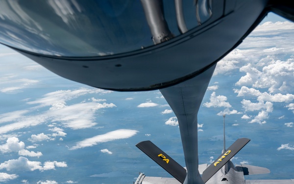 An aerial refueling operation is captured from the perspective of a tanker aircraft, showing its refueling boom extended toward an F-16 Fighting Falcon fighter jet. The F-16 is marked with 'PA' and 'ANG' in yellow lettering, indicating it belongs to the Pennsylvania Air National Guard. The aircraft are flying at altitude above a partially cloud-covered landscape.